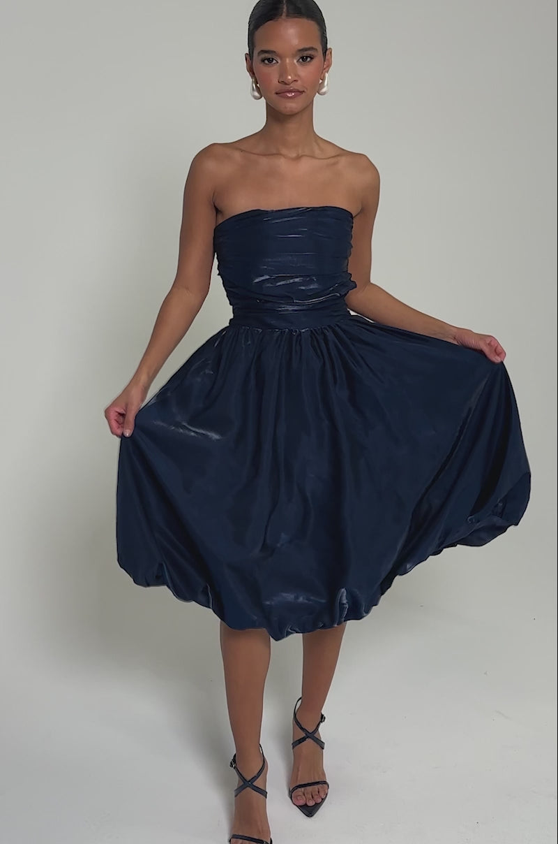 Video of woman modeling the LUXE LIFE SATIN MIDI DRESS, a strapless navy blue midi featuring a ruched bodice and voluminous shiny skirt. Her hair is pulled back and she accessorizes with large hoop earrings.