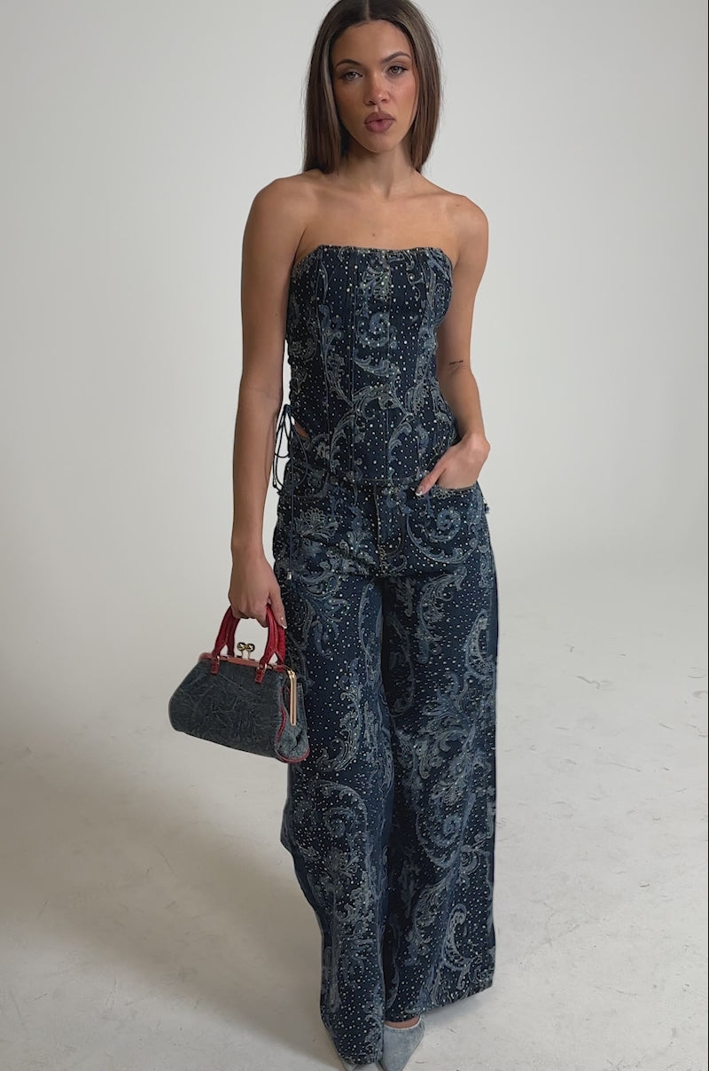 Video of woman modeling the ELEVATED DENIM CORSET WITH RHINESTONES and matching pants, holding a red and blue bag in her left hand.
