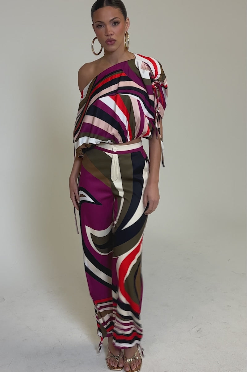 Video of woman modeling the CRAZY IN LOVE TROUSER—retro-chic, high-rise wide-leg pants in pink, brown, and cream—paired with a colorful off-shoulder top, gold heels, and large hoop earrings against a plain background.
