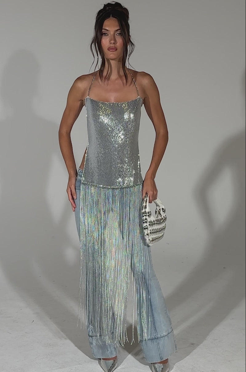A video of a woman posing in the ONLY THINK ABOUT YOU SILVER RHINESTONE FRINGE TOP featuring a shiny sequin embellished bodice and halter neckline, paired with light blue flared pants, silver heels, and a white clutch against a plain background.
