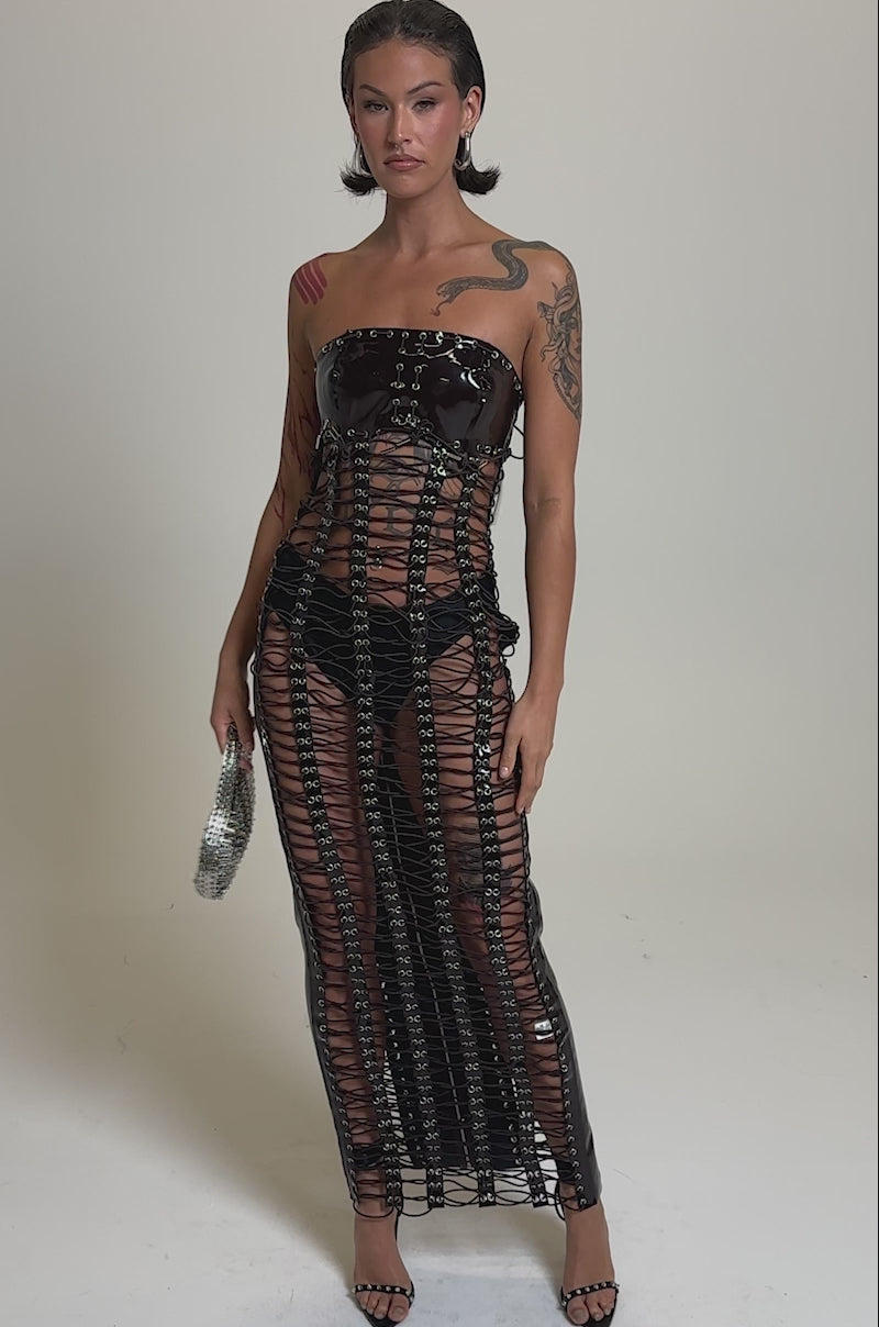 A video of a woman with wet-look hair and tattoos stands confidently against a plain, light background wearing the ONE OF ONE LACE UP MAXI DRESS, featuring see-through chainlink details over black underwear, and holding a small handbag.