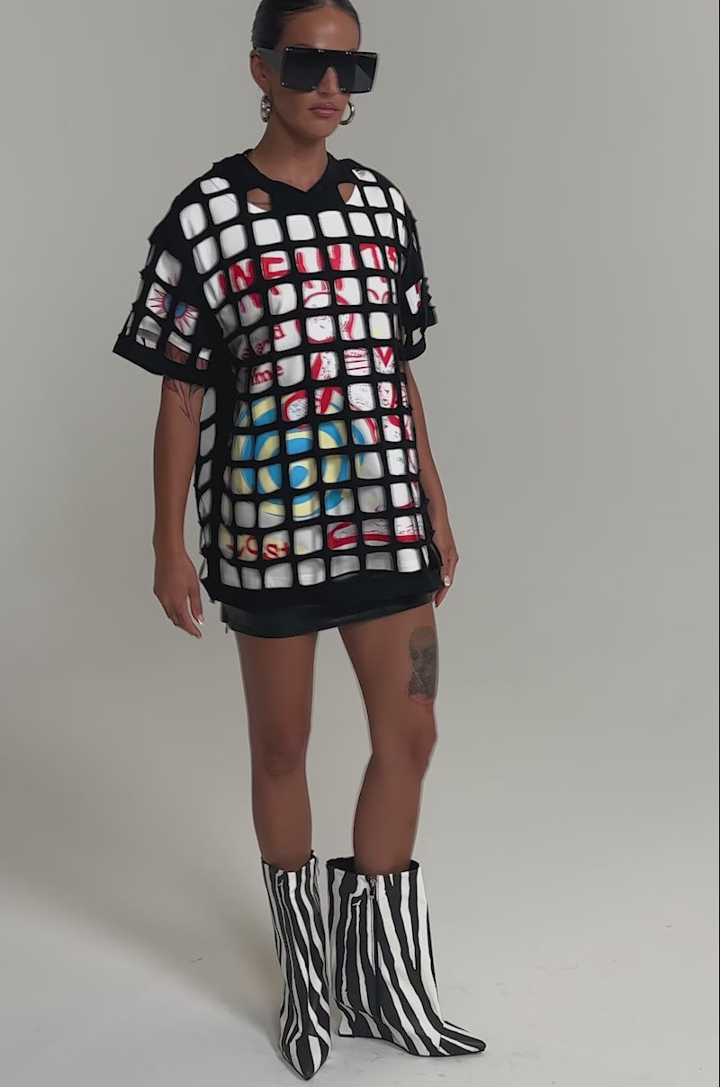 Video of woman modeling the CHECKED OUT OVERSIZED TEE layered under a black mesh top with a black leather mini skirt, creating a bold streetwear look against a plain background.