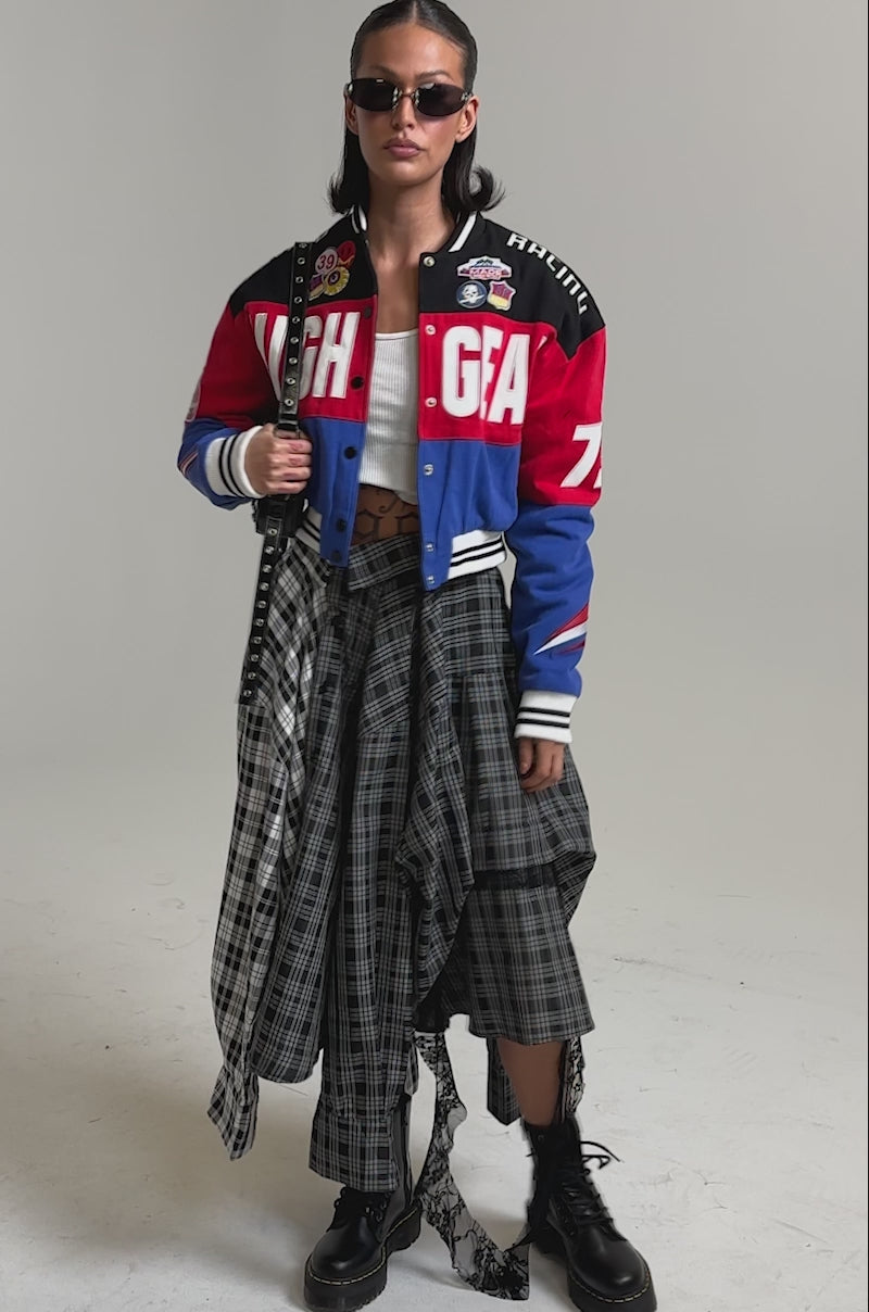 A video of a woman wearing sunglasses, chunky black boots, a long plaid skirt, black belt, and the HIGH GEAR EMBROIDERED BOMBER with colorblock style and racing-inspired details.