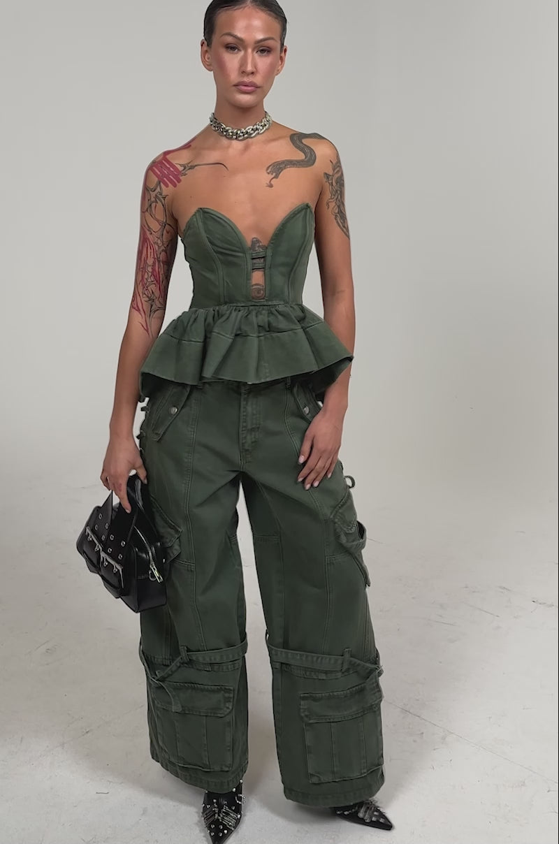 Video of woman modeling the KARA DENIM CORSET and matching pants in olive green, showing her left arm tattoos and styling a bold silver statement necklace against a plain light gray background.