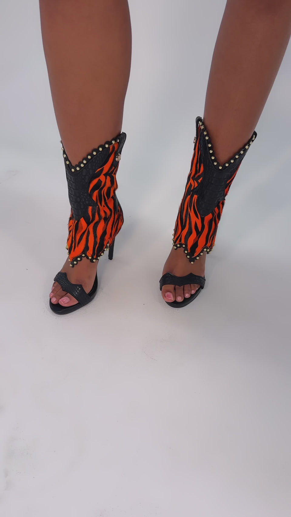 A video of a person wearing the AZALEA WANG TAHLIE ORANGE MULTI WESTERN GLADIATOR SANDAL with bold orange and black tiger-striped, gold-studded leg accessories, standing on a white background.