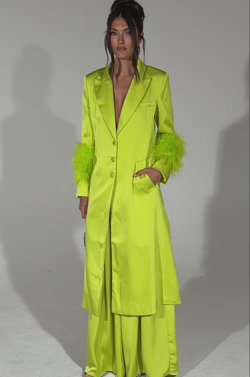 A video of a woman posing against a plain light background wearing the CHARTREUSE FEATHER TRIM TRENCH over matching wide-leg pants and holding a small blue textured handbag.