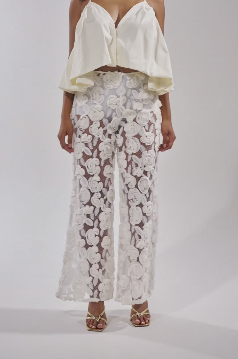 A video of a woman wearing the DINA HALTER NECK BLOUSE in ivory poplin, styled with sheer flared pants featuring white floral embroidery. 