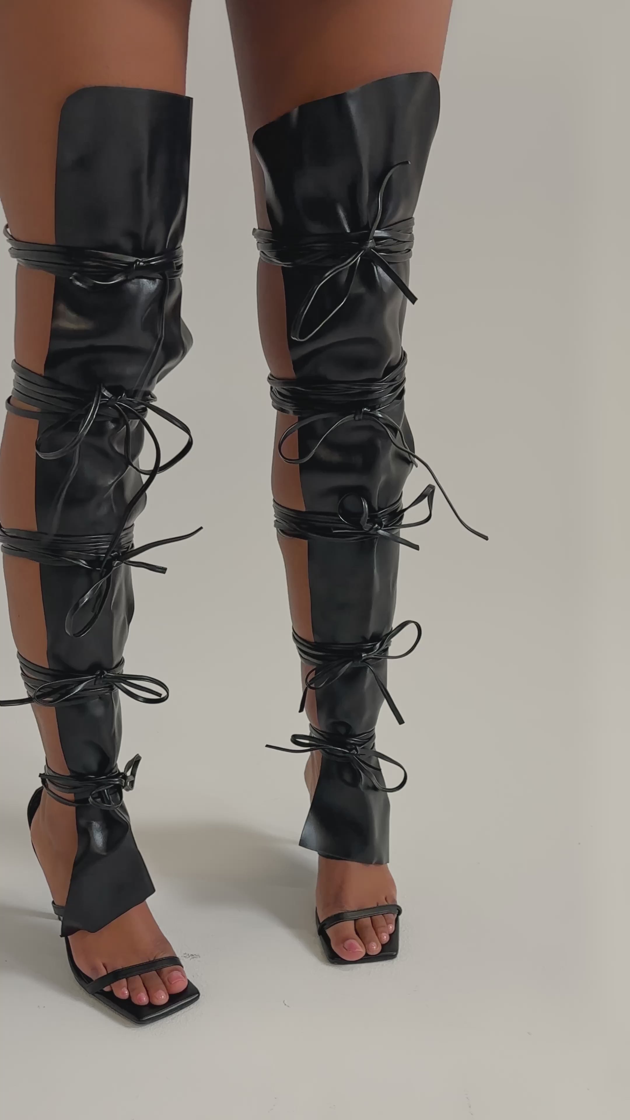 A video of a person wearing the AZALEA WANG KRISTINE BLACK STRAPPY UPPER GLADIATOR SANDALS, featuring thigh-high lace-up details with large cutouts and stiletto heels.