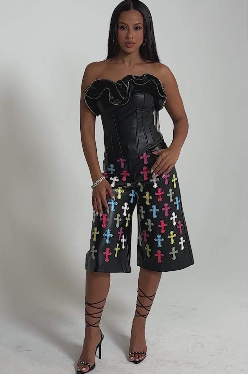 A video of a woman wearing the DON'T MESS RUFFLE CORSET, a black strapless top with ruffled zippered details, paired with matching knee-length shorts featuring colorful cross patterns. 
