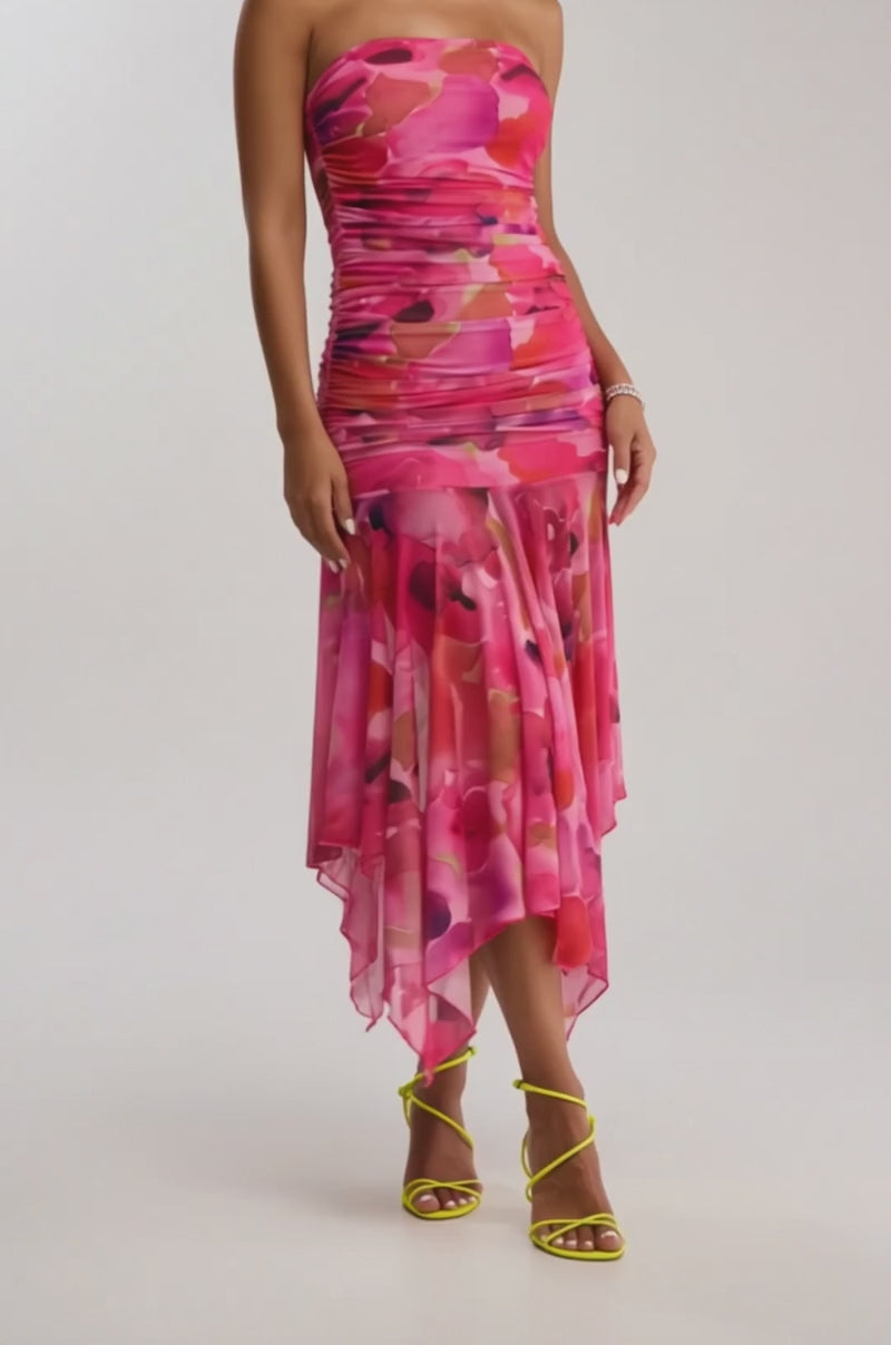 A video of a woman in the BRUNCH DATE MESH MIDI DRESS IN PINK MULTI, featuring pink ruched mesh with red florals and an asymmetrical hem, paired with yellow strappy heels against a plain light background.
