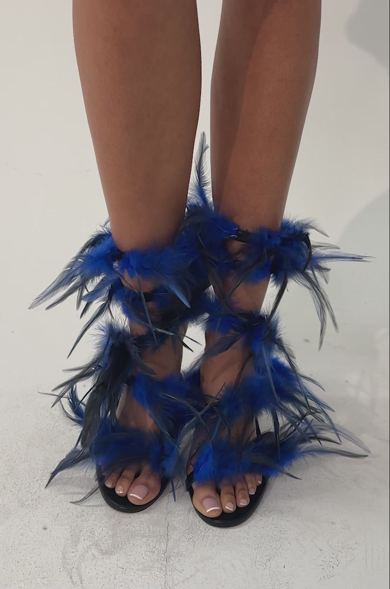 A video of a person wearing the AKIRA GOLD COVERT FEATHER EMBELLISHED SPIRAL STRAP STILETTO SANDALS against a neutral background.