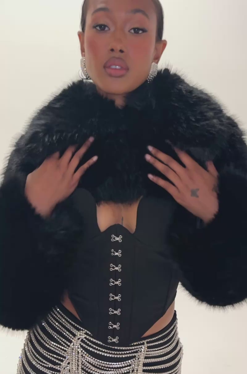 Video of a woman modeling the LEILA ULTRA CROP FAUX FUR IN BLACK, pairing the black cropped faux fur jacket with a black corset featuring hook detailing.