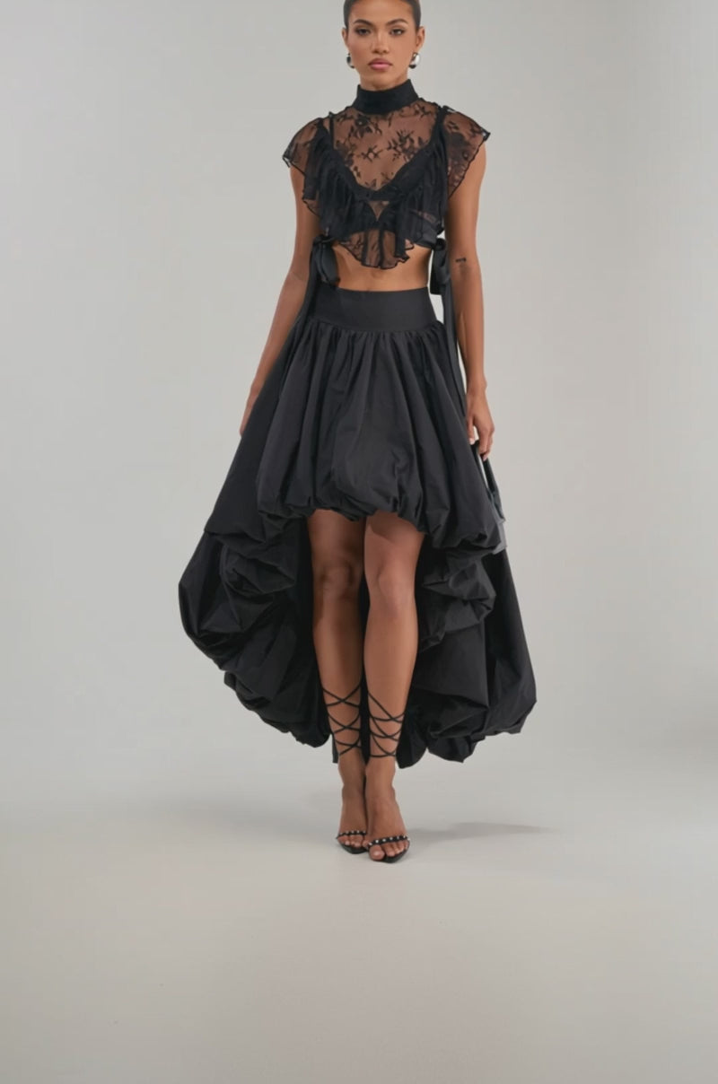 Video of woman modeling the THE BIGGER THE BETTER HIGH LOW MAXI SKIRT IN BLACK, styled with a small black handbag and strappy black heels, standing against a plain background.