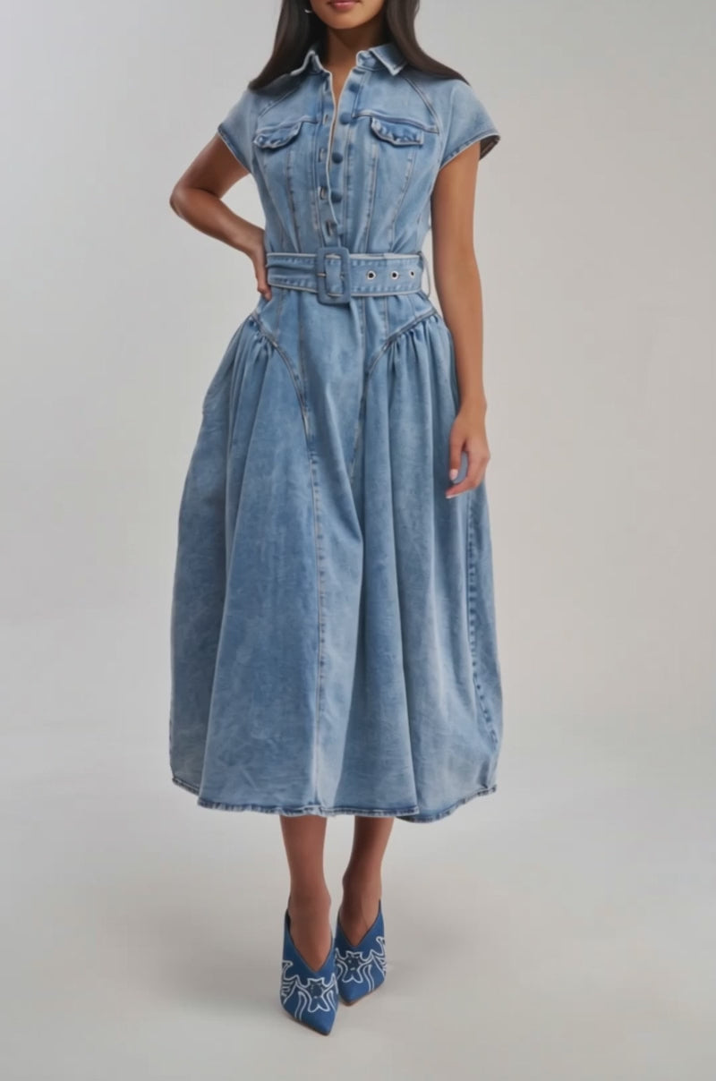 Video of woman modeling the MEET THE PARENTS BELTED DENIM MIDI DRESS, featuring a collar, short sleeves, front pockets, pleats, and a belt. She pairs it with blue heels with white patterns against a plain light background.