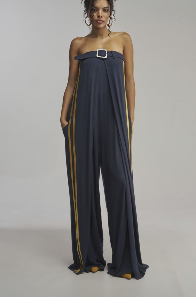 Video of woman modeling the RUN THE SHOW JUMPSUIT IN NAVY—featuring a strapless wide-leg silhouette, bold yellow side stripes, a belted waist with a silver buckle, and paired with yellow shoes