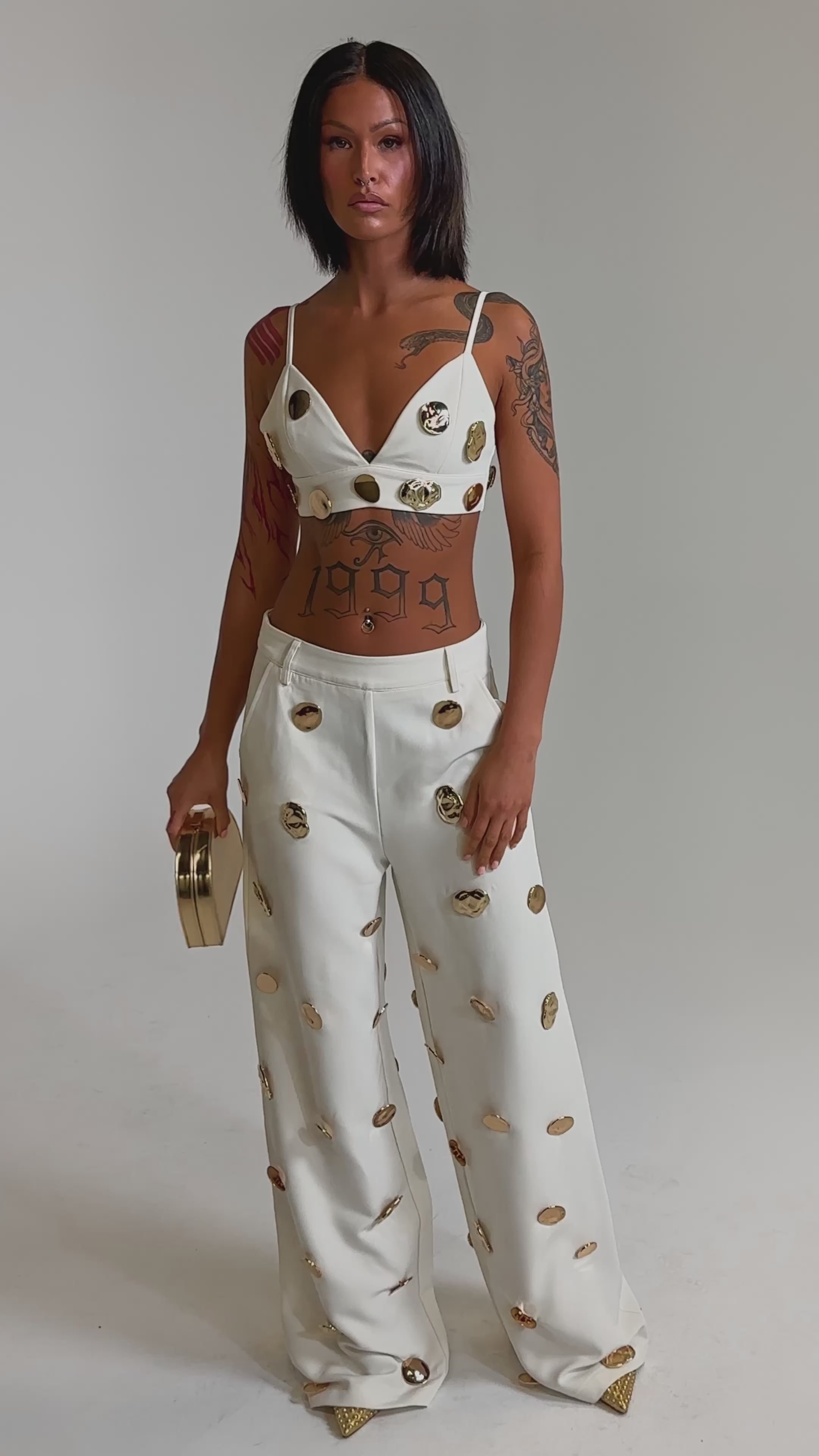 A video of a woman wearing the SARASOTA EMBELLISHED BRALETTE and wide-leg pants with gold-tone disc details. She holds a metallic clutch, sports pointed gold shoes, and poses confidently against a plain background.