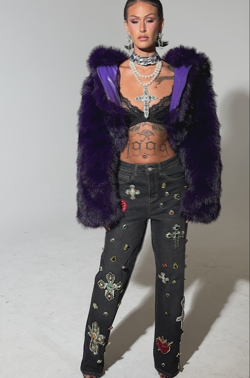 A video of a woman wearing the JUST A FLING CROPPED FAUX FUR jacket with a hood, jeweled bralette, statement necklace, and high-waisted black jeans decorated with colorful gems and cross patches.