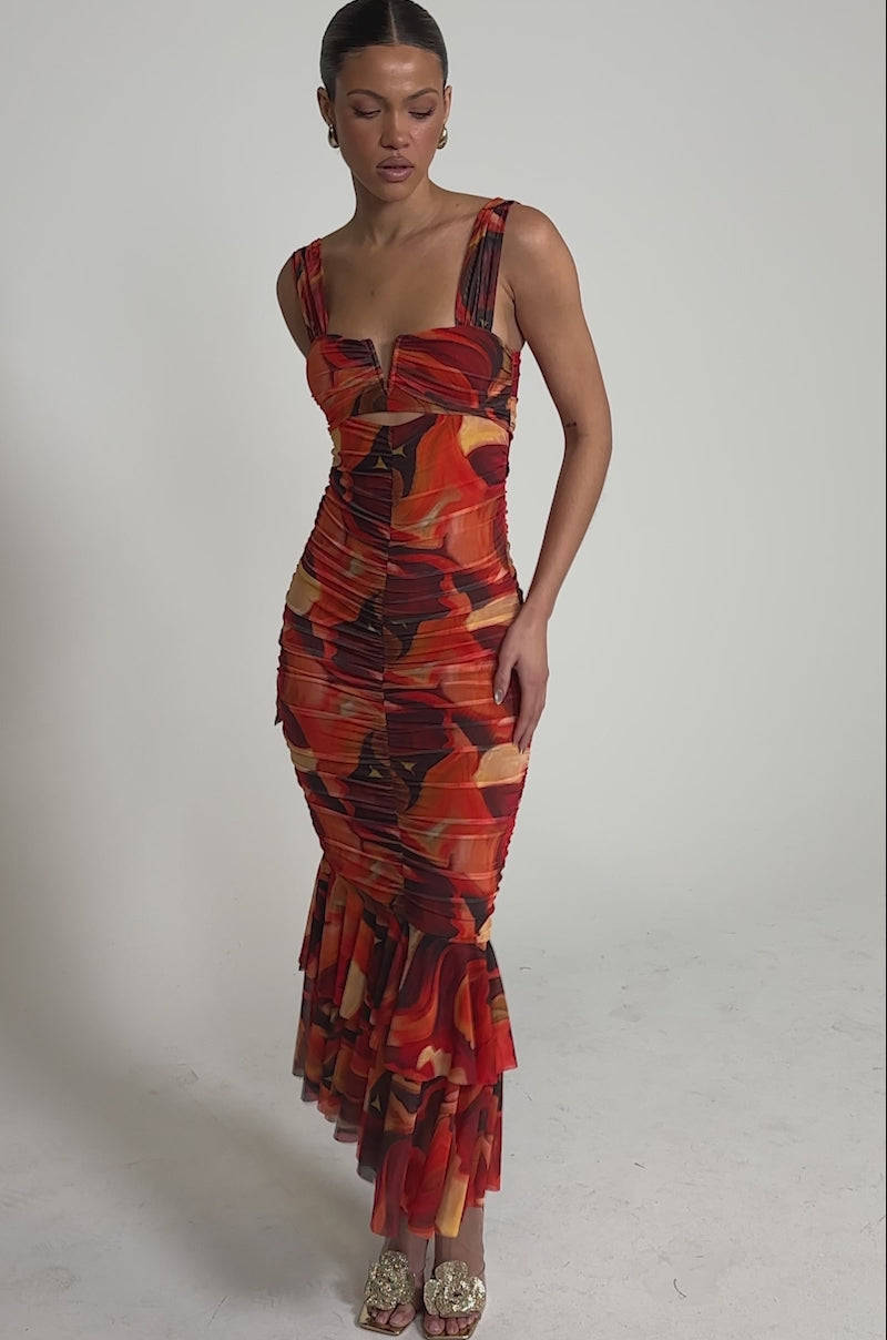 A video of a woman in the BELONG IN A MUSEUM RED MAXI DRESS, featuring a form-fitting silhouette, sleeveless design, flame print, and flared hem, standing against a light background with open-toe heels.