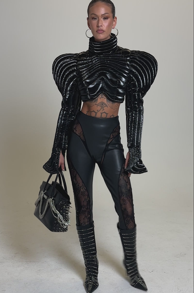 A video of a woman posing in LACE DETAIL PANELED FAUX LEATHER LEGGINGS IN BLACK, paired with a futuristic black top, studded boots, hoop earrings, and a structured chain handbag. Her hair is pulled back, highlighting the leggings' lace details and bold style.