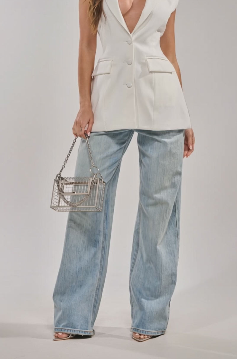 A video of a woman with wavy brown hair poses confidently in the HARLOW BACKLESS BLAZER VEST in white, paired with clear heels and denim pants, against a plain white background.