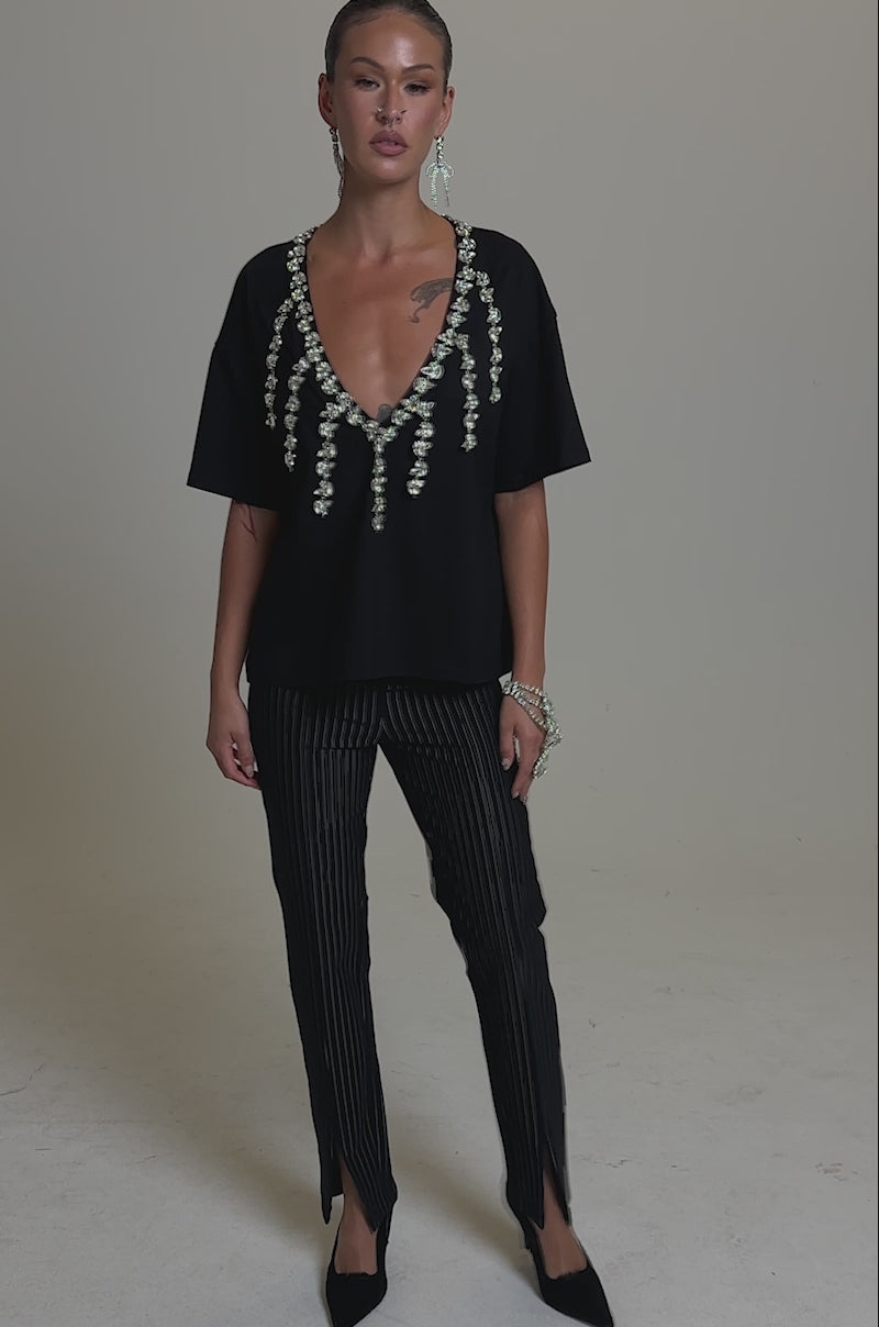 A video of a person in the RHINESTONE QUEEN OVERSIZED TEE with a plunging V-neckline and hanging embellishments, paired with black pinstripe pants with front slits, black heels, and statement earrings against a plain background.