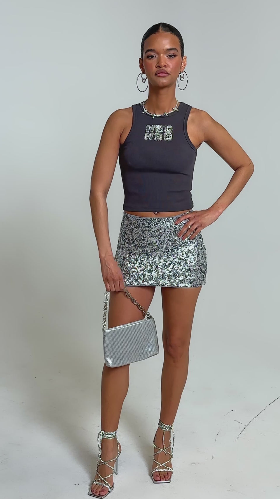 Video of woman modeling the NO BIG DEAL TANK—a dark ribbed style with rhinestone lettering—paired with a silver sequin skirt, hoop earrings, a necklace, and her hair pulled back for a Y2K-inspired look.