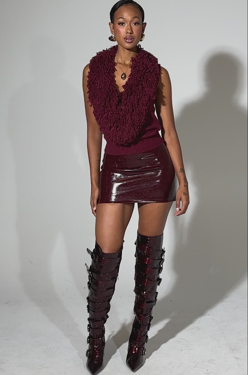 A video of a woman wearing the AKIRA Label CARLY FRINGE OPEN FRONT SWEATER in burgundy, paired with a shiny burgundy mini skirt and tall, dark burgundy buckle boots.