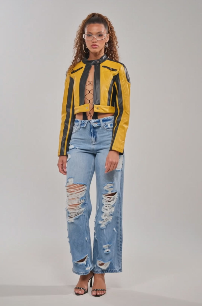 Video of woman modeling the ON YOUR MARKS RACER MOTO IN YELLOW MULTI jacket, a cropped jacket paired with a lace-up black top and ripped high-waisted blue jeans. She has curly hair in a ponytail and clear fashion glasses for a bold biker look.