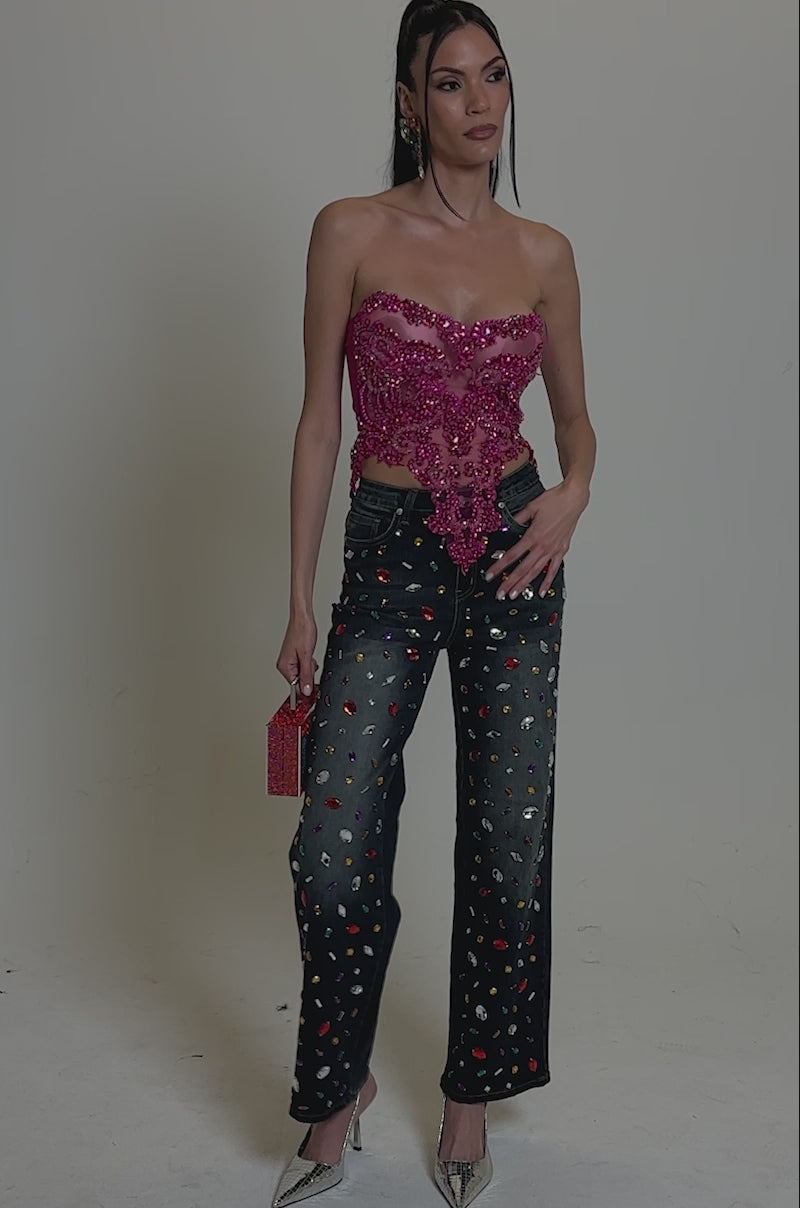 Video of woman modeling the JEWELERS ROW RHINESTONE DENIM JEANS, covered with colorful gems, paired with a sparkly pink top, silver heels, and a small glittery purse is shown.