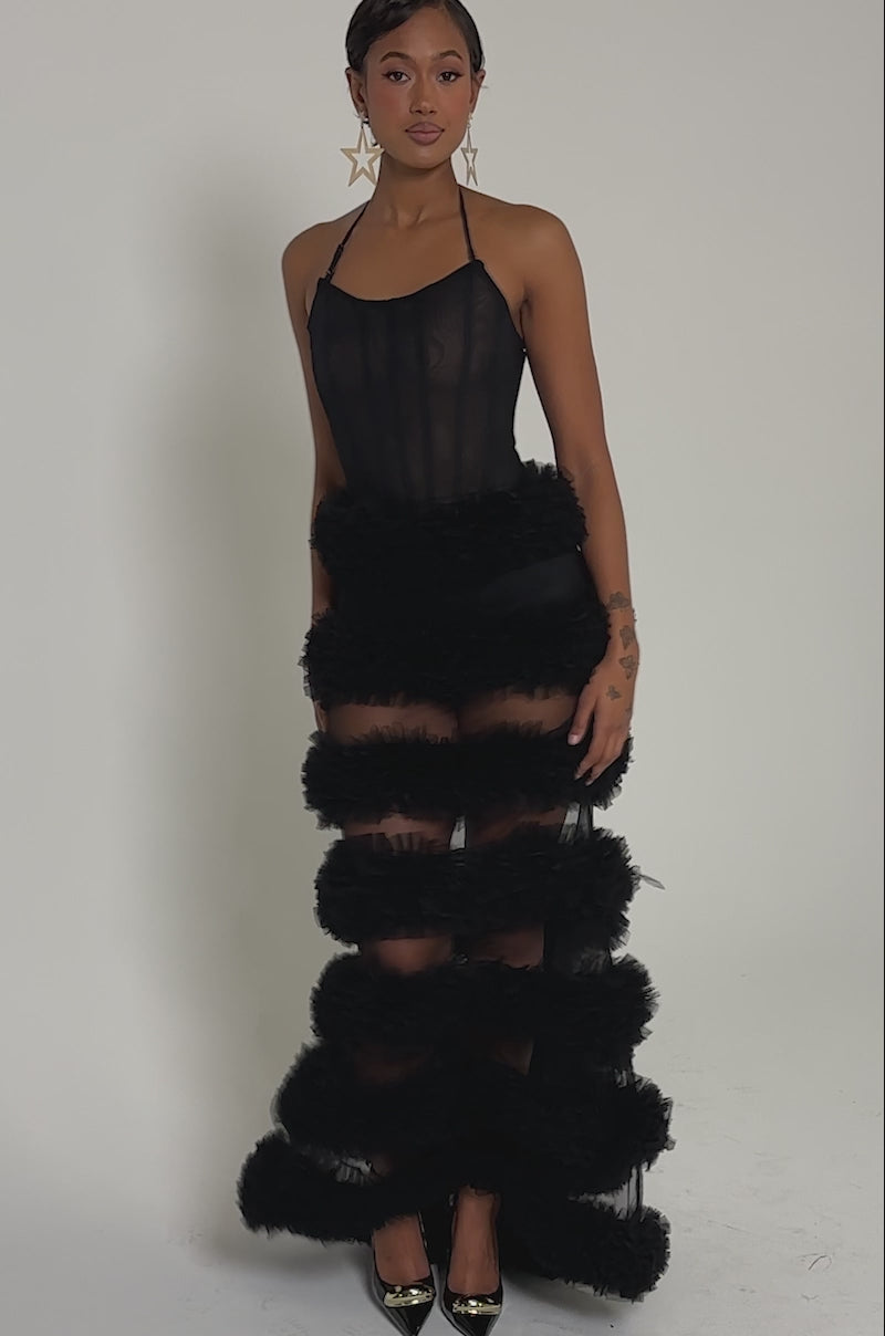 Video of woman modeling the QUEEN OF EVERYTHING MAXI DRESS, a black sheer tulle maxi with horizontal ruffles from waist to hem, styled with gold pointed shoes and dangling earrings, posing against a plain light background.