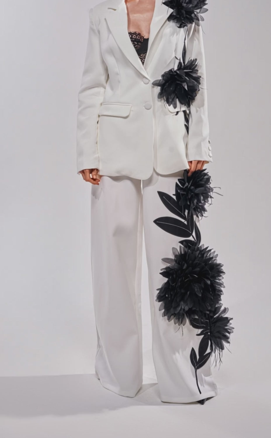 A video of a woman wearing the IN FULL BLOOM APPLIQUE TROUSER, a white wide-leg suit with oversized black floral embroidery on the jacket and trousers. 