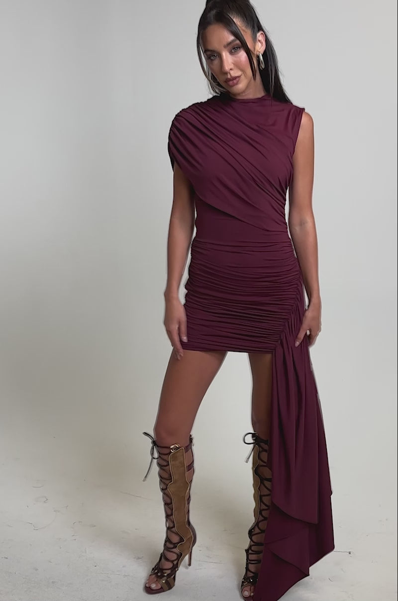 A video of a woman in the WRAP ME UP MINI DRESS, a sleeveless burgundy wrap mini with ruching and an asymmetrical draped panel, paired with lace-up knee-high heels.