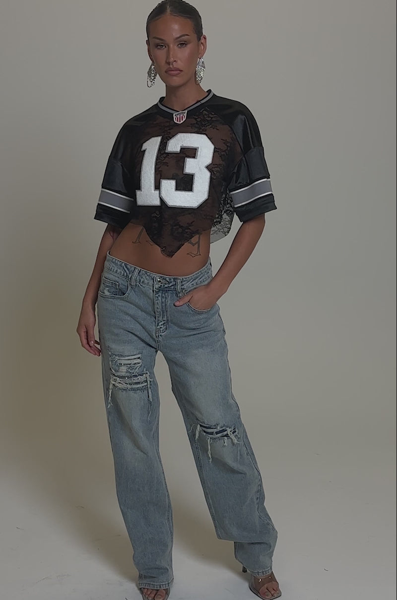 Video of woman modeling the RUN AWAY WITH ME RELAXED DENIM ripped jeans, a cropped black lace football jersey with number 13, hoop earrings, and open-toed heels against a plain gray background.