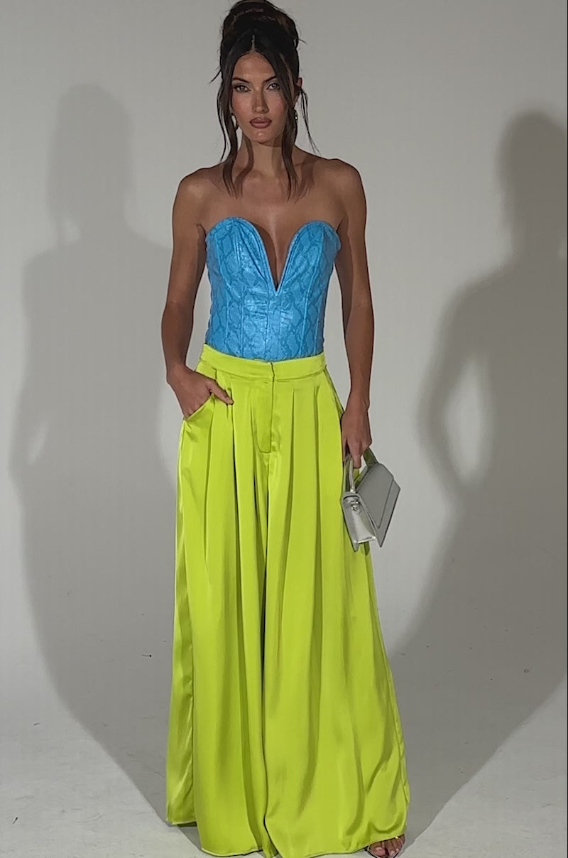 A video of a woman posing in a turquoise strapless corset top and CHARTREUSE PLEATED PALAZZO PANT with a wide-leg, pleated design, holding a small silver handbag against a plain light background.