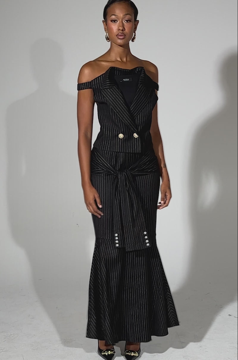 A video of a woman modeling the BUSINESS ONLY PINSTRIPE CORSET, a black strapless pinstripe dress with a fitted waist, flared skirt, and decorative buttons. She pairs it with black pointed heels and wears her hair slicked back against a light gray background.