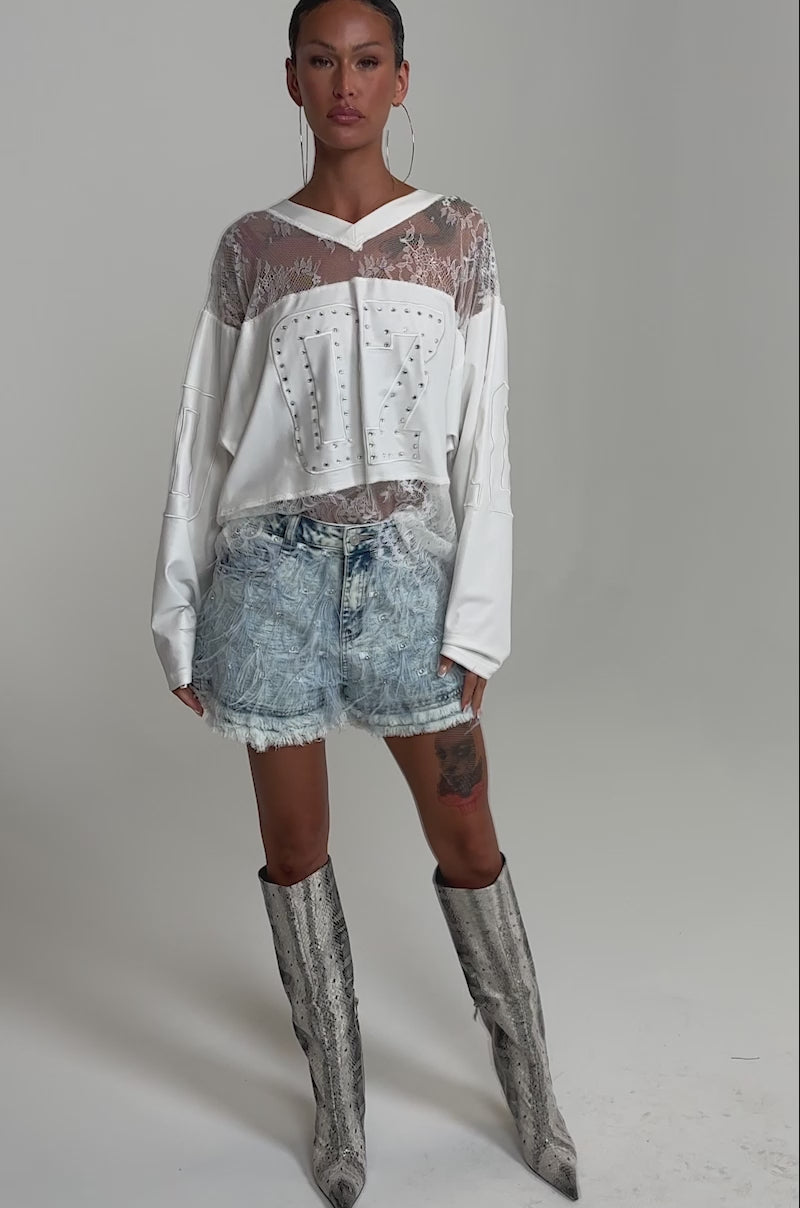 A video of a woman in the CHECK IT OUT BABY DENIM SHORT, paired with a sheer white embroidered top and knee-high snakeskin boots, accessorized with large hoop earrings.