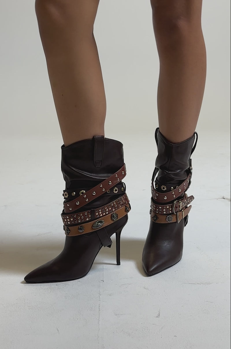 A video of a person wearing the AZALEA WANG BROWN STILETTO BOOTIE: dark brown pointed-toe heels featuring multiple leather straps, metal studs, conchos, and a bold gold buckle.
