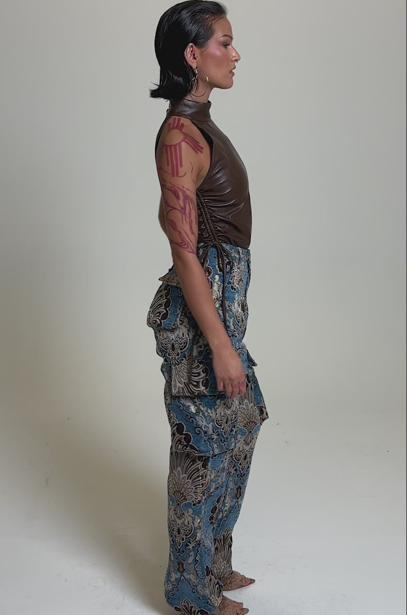 A video of a person wearing a sleeveless brown leather halter top and blue high rise THE DUOMO PANT with a paisley design, paired with open-toe heels, against a plain light background.