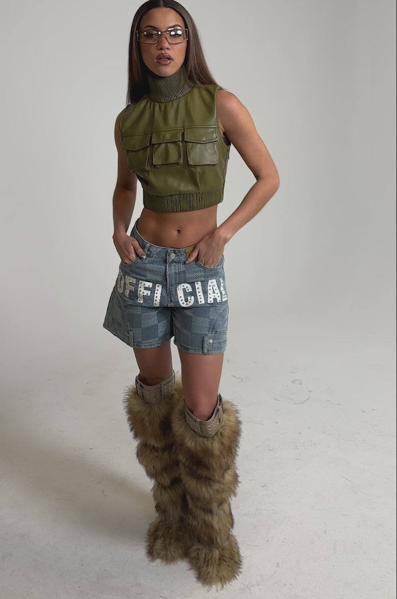 Video of woman modeling the VANJA MOCKNECK FAUX LEATHER TOP—an olive, sleeveless, cropped mock neck with four front pockets—standing with her hands on her hips, paired with blue jean shorts featuring white lettering and checkered patterns.