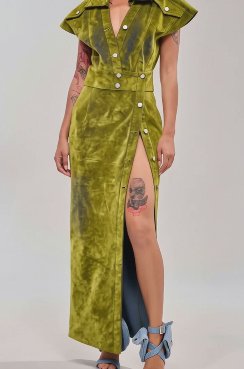 Video of woman modeling the CHELSEA BUTTON DOWN MAXI DRESS IN CHARTREUSE VELVET DENIM, featuring a sleeveless design, deep V-neck, and thigh-high slit that reveals her skull tattoo. She pairs it with blue heeled sandals with large bows and has her hair pulled back.