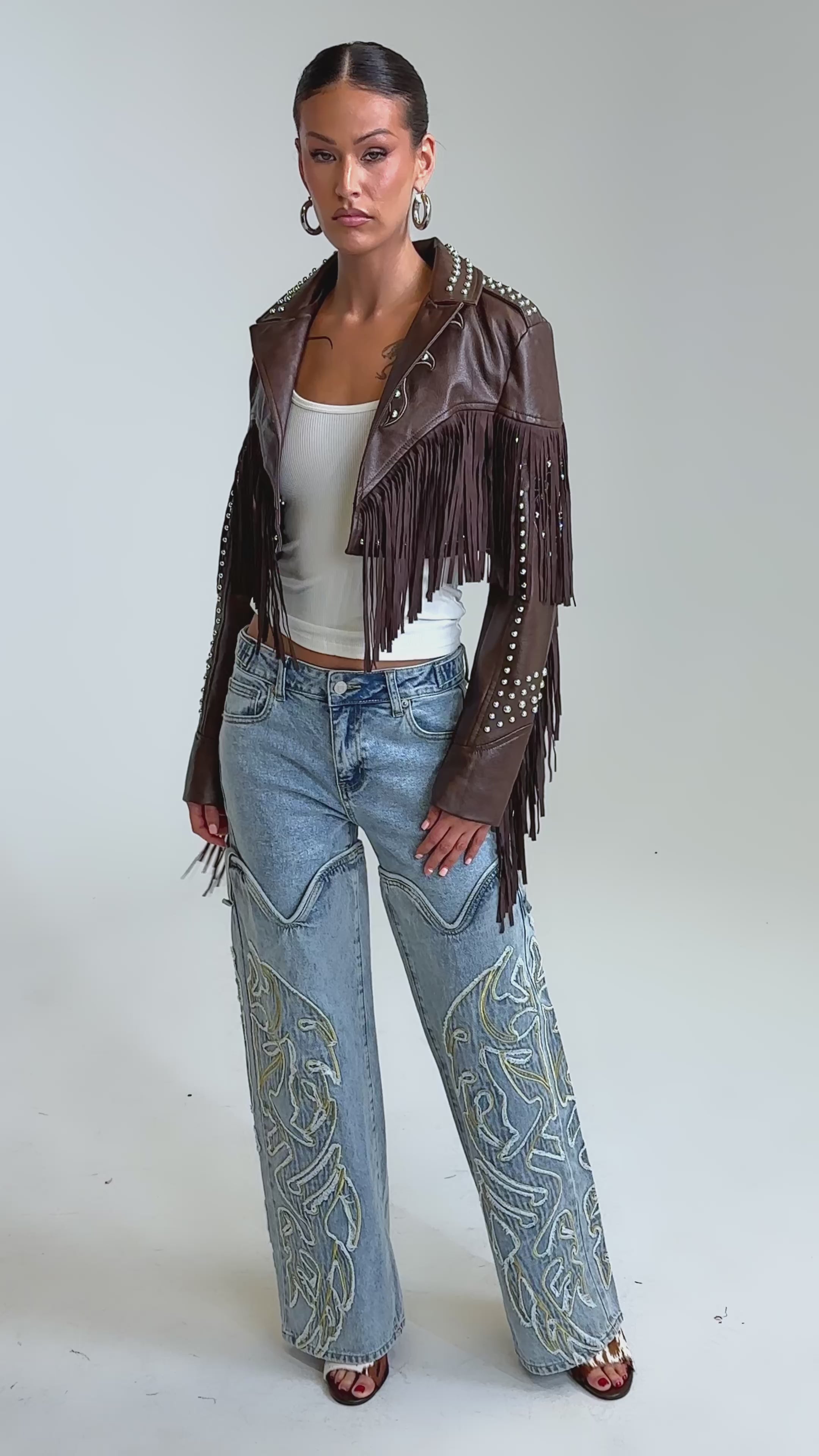 Video of woman modeling the NOT MY FIRST RODEO CROPPED MOTO—a vintage-inspired western faux leather jacket with fringe and silver studs—pairs it with a white top, large hoop earrings, and light blue jeans, standing confidently against a plain backdrop.