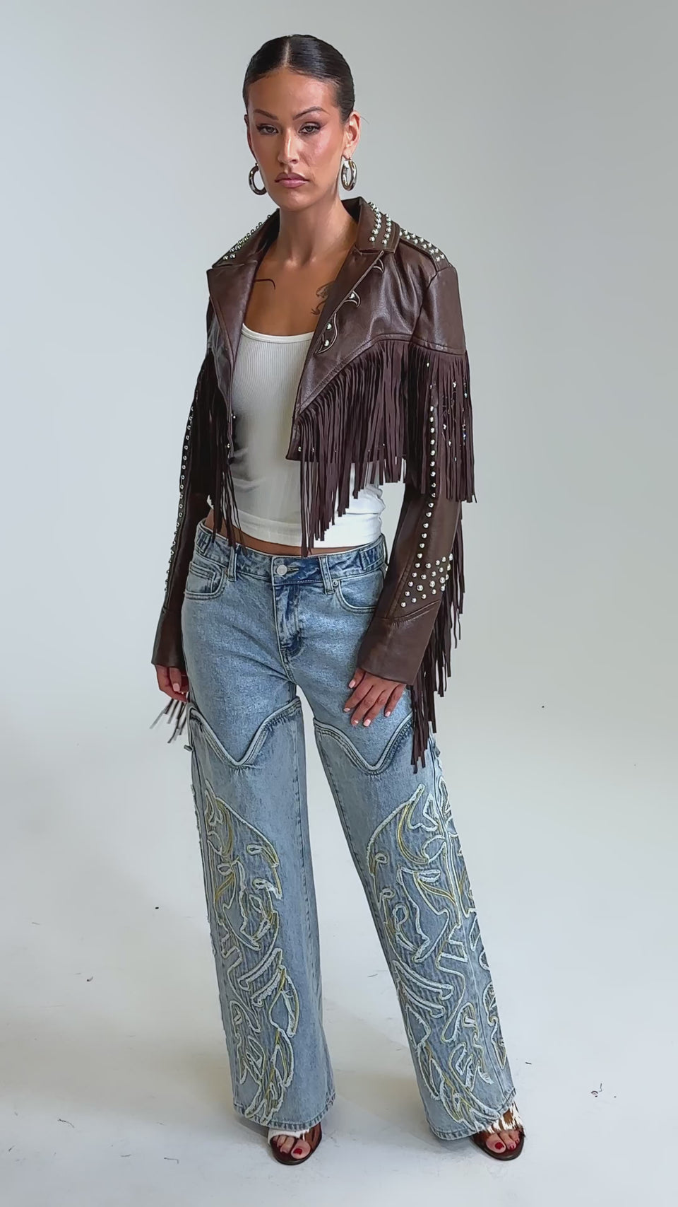 Video of woman modeling the NOT MY FIRST RODEO CROPPED MOTO—a vintage-inspired western faux leather jacket with fringe and silver studs—pairs it with a white top, large hoop earrings, and light blue jeans, standing confidently against a plain backdrop.