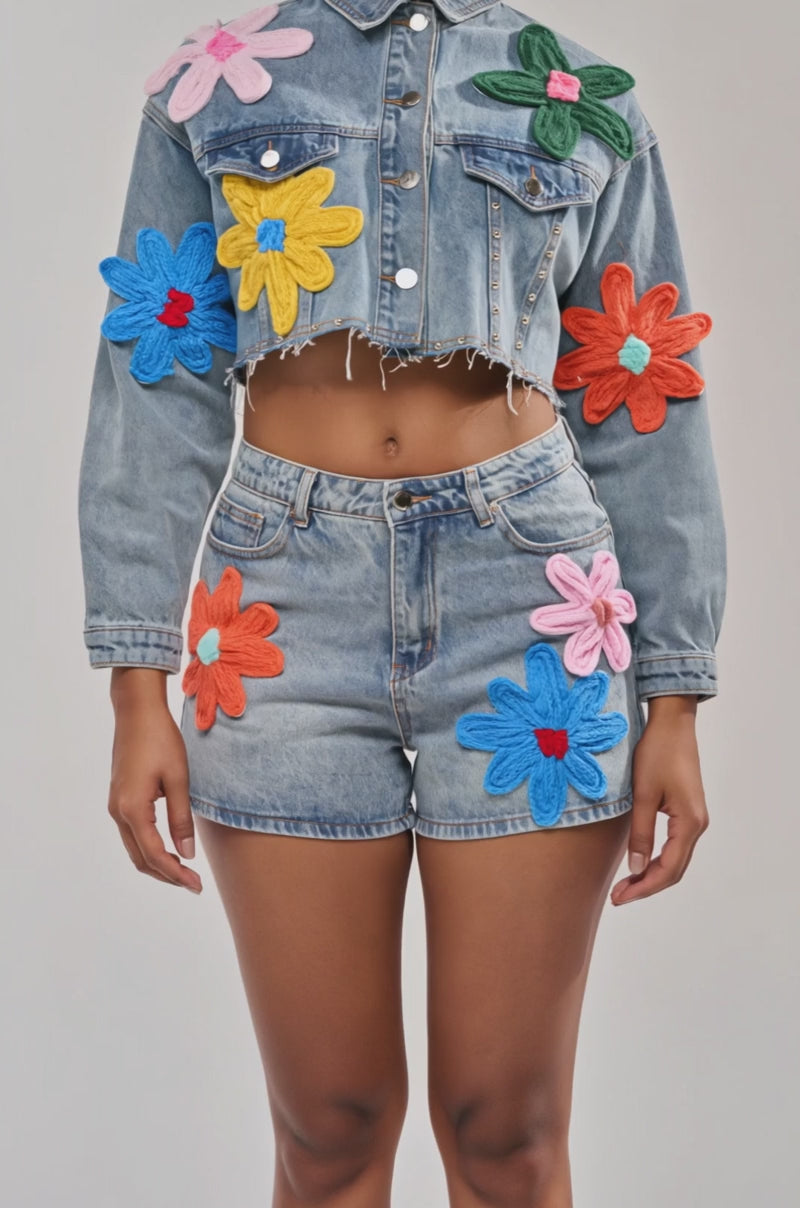 Video of woman modeling the FLOWER PATCH DENIM JACKET, featuring large, colorful floral appliques and a cropped distressed hem, paired with matching shorts adorned with bright flower patches, against a plain background.