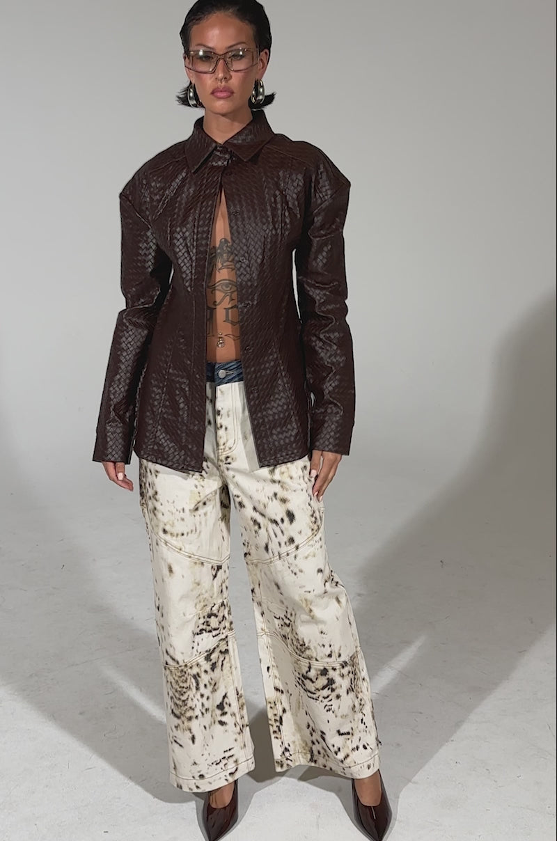 A video of a person wearing the FEELING RICH FAUX LEATHER BUTTON DOWN—a textured dark brown jacket with a deep neckline and adjustable buckled belt—paired with wide-leg abstract print pants, brown heels, clear glasses, and displays chest tattoos against a plain background.