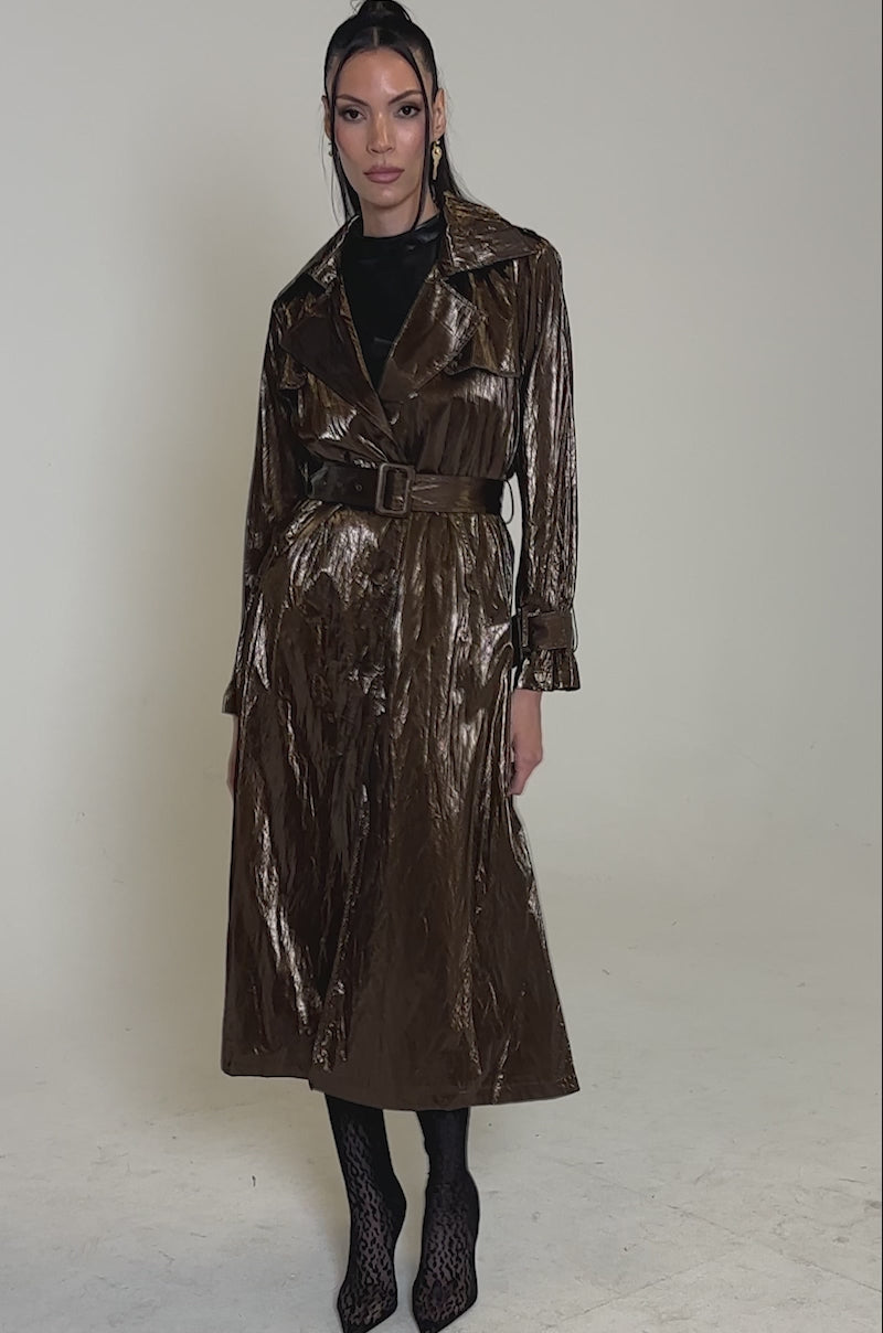 A video of a woman with long dark hair striking a confident pose against a plain background, wearing the WORK OF ART METALLIC TRENCH in bronze over a black outfit, paired with knee-high patterned boots.