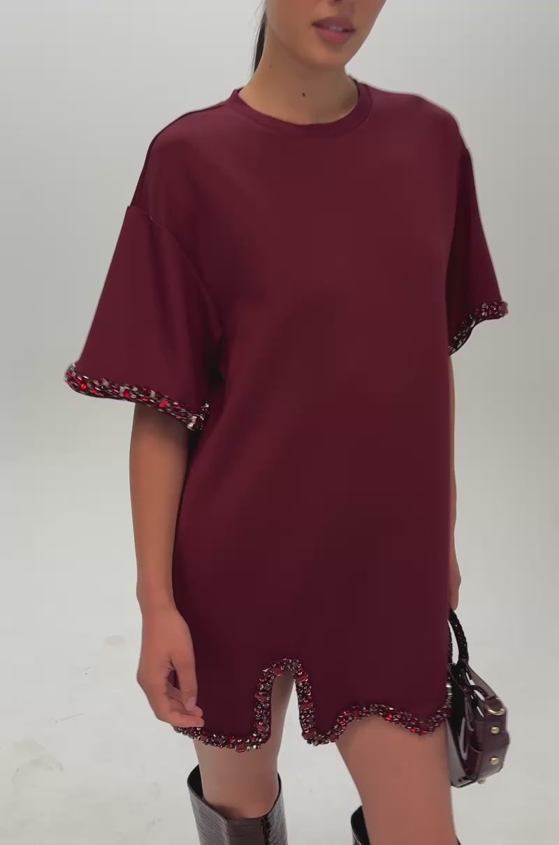 Video of a woman modeling the COOL GIRL EMBELLISHED TEE SHIRT DRESS IN BURGUNDY, a rhinestone-adorned dress featuring short sleeves, a high-low hem, and a side slit. The sleeve and hem edges are embellished with dark red faceted crystals for extra sparkle.