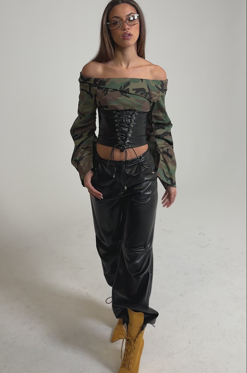 Video of woman modeling the TIME TO FIGHT LACE UP CAMO CORSET BLOUSE with flared sleeves, paired with sleek leather pants and sunglasses against a plain light gray background.