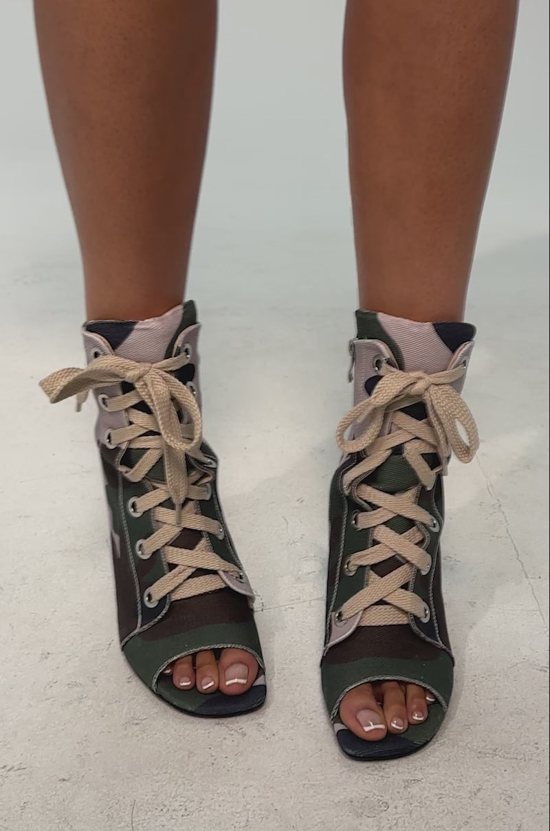A video of a model wearing the AZALEA WANG ROOFTOP CAMO OPEN TOE LACE UP BOOTIE. It is a high-heeled stiletto bootie with open toes, canvas construction, and a green, brown, tan, and black camouflage print.