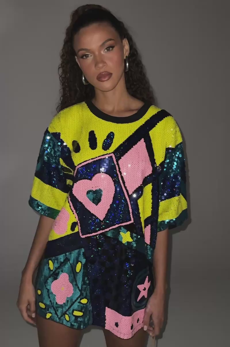 Video of woman modeling the QUEEN OF HEARTS SEQUIN TSHIRT DRESS, an oversized piece with multicolor geometric patterns, hearts, stars, and shades of blue, yellow, pink, and black against a plain background.