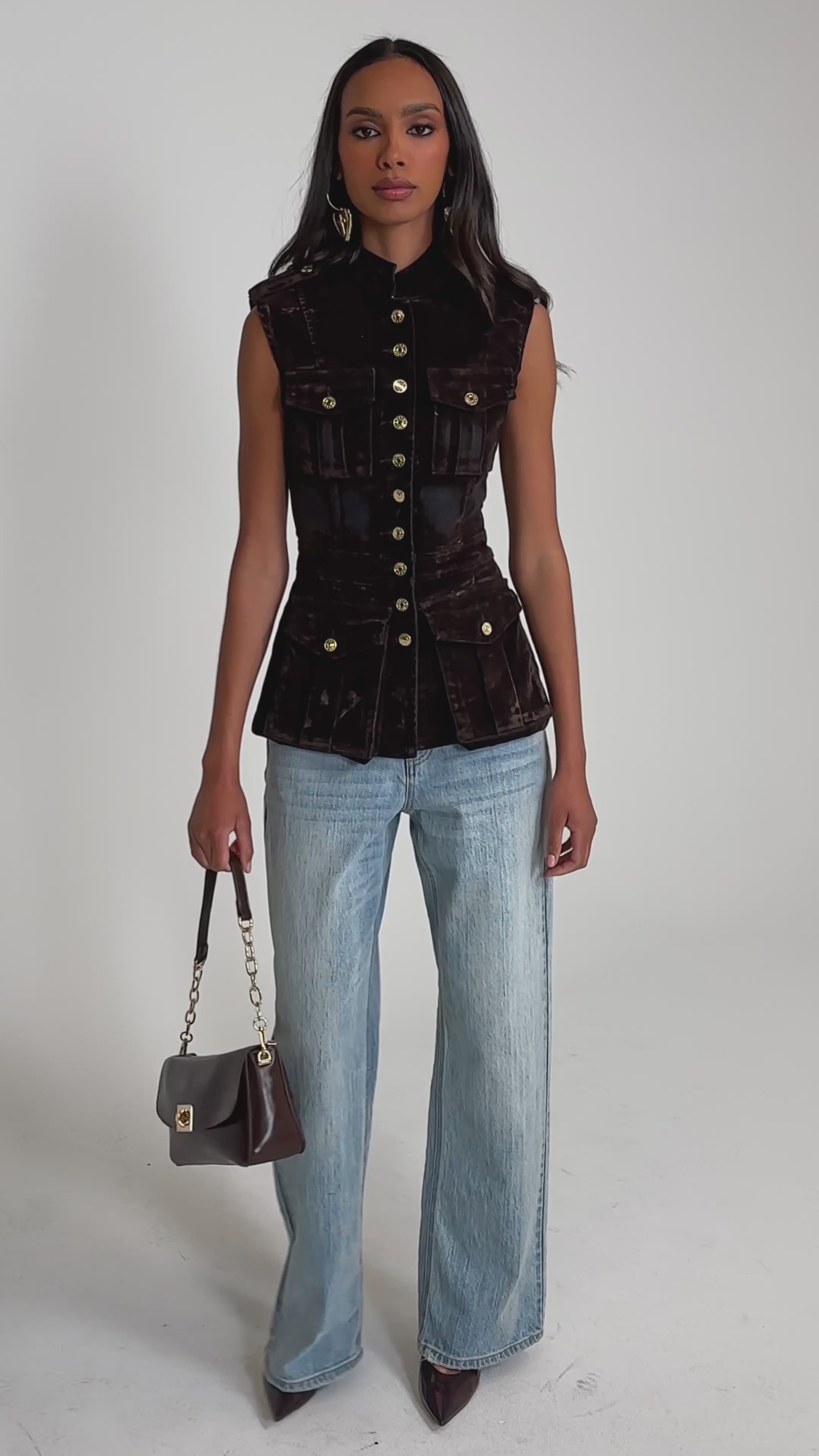 Video of woman modeling the BACK WITH IT VELVET BUTTON DOWN, a fitted brown military-style vest with gold buttons and flap pockets, paired with light blue jeans against a plain light gray background.